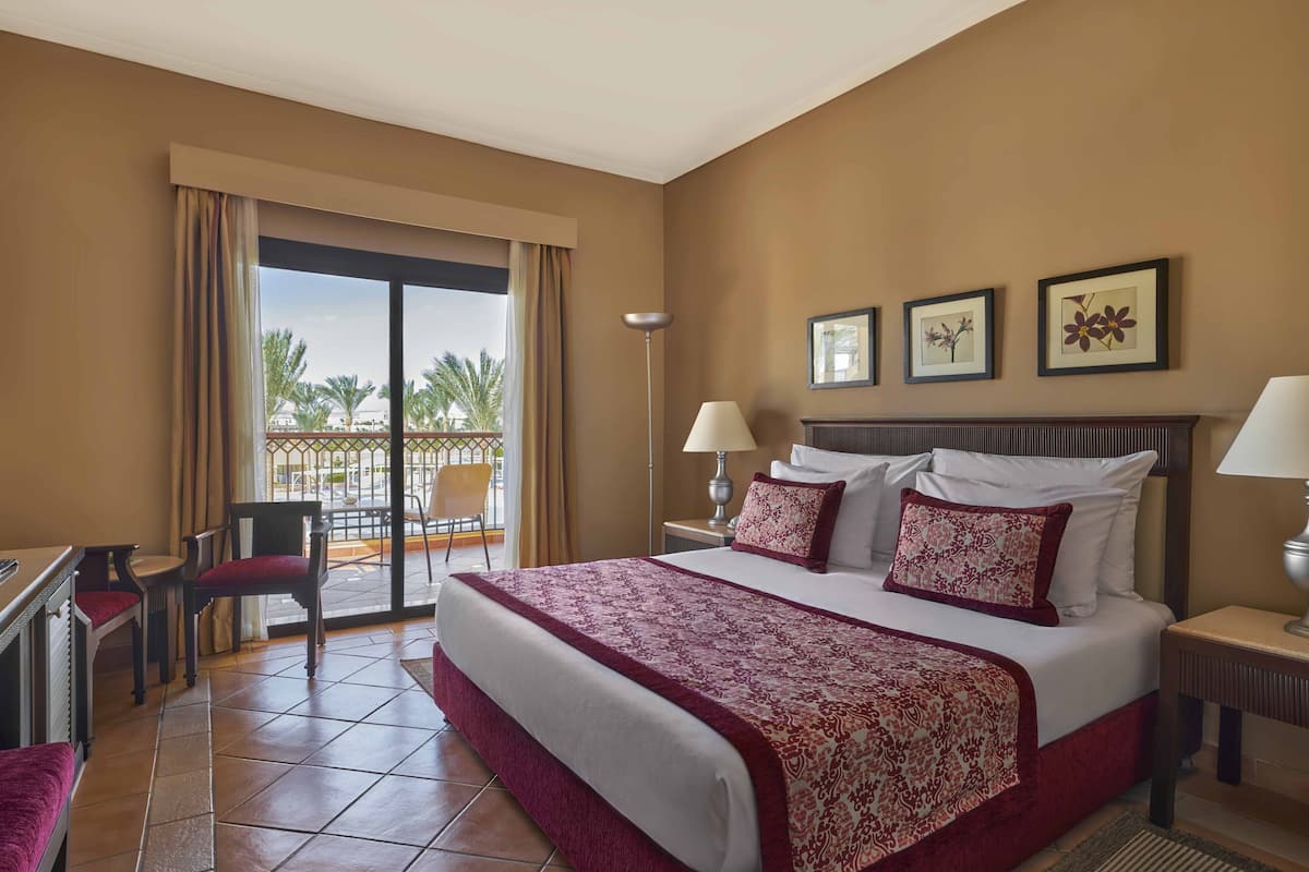A hotel room with a large bed, a desk, and a chair. There is a balcony with a view of palm trees.