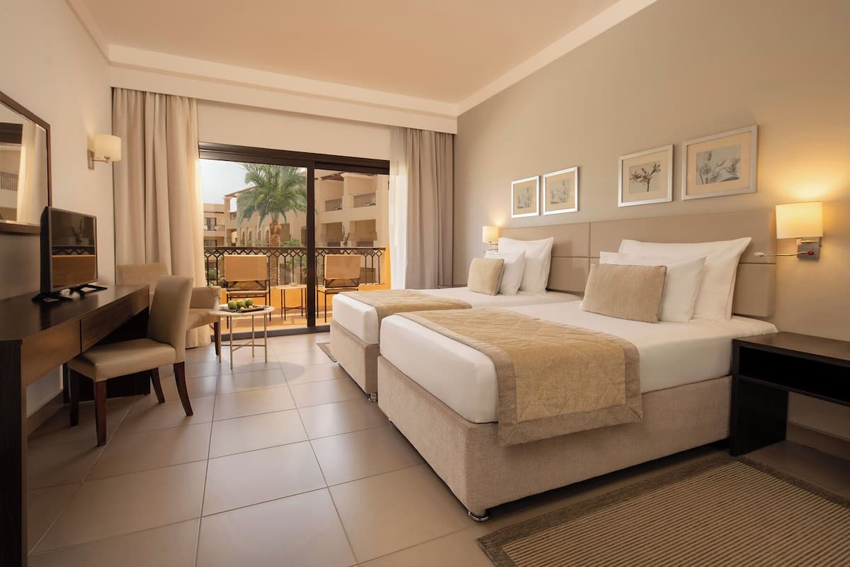 A hotel room with two beds, a desk, a chair, a TV, and a balcony with a view of palm trees.