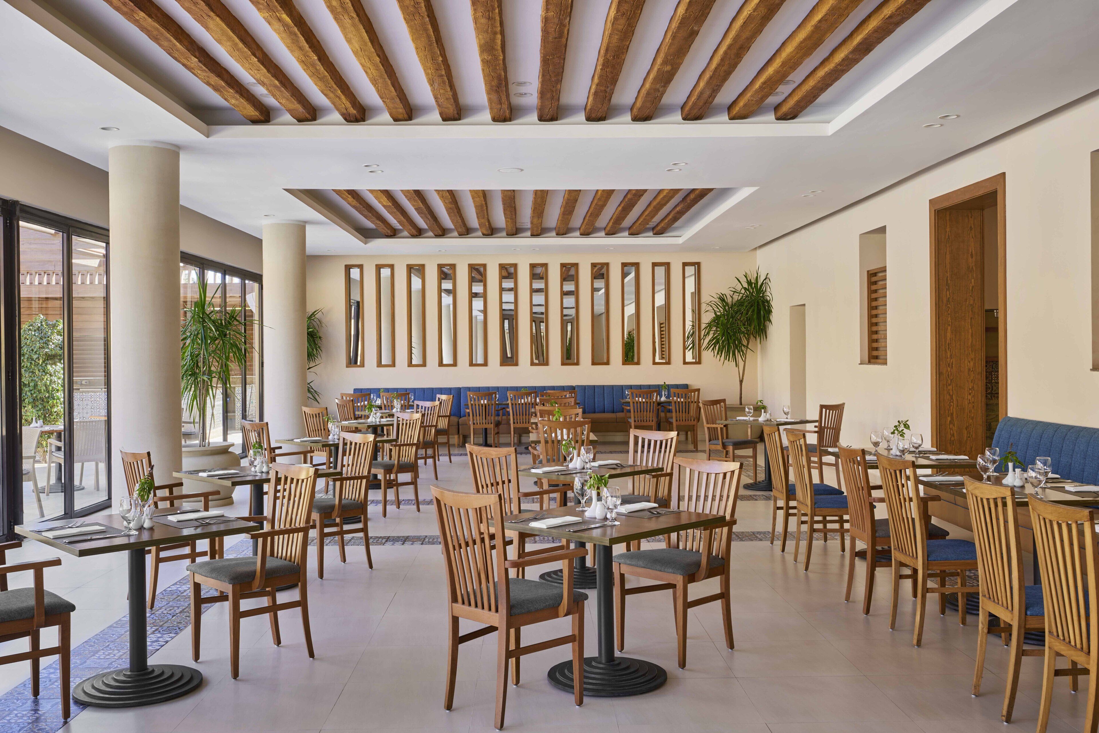 Un vaste espace dédié à la salle à manger, avec des tables et des chaises en bois, de grandes fenêtres et un plafond haut avec des poutres apparentes.