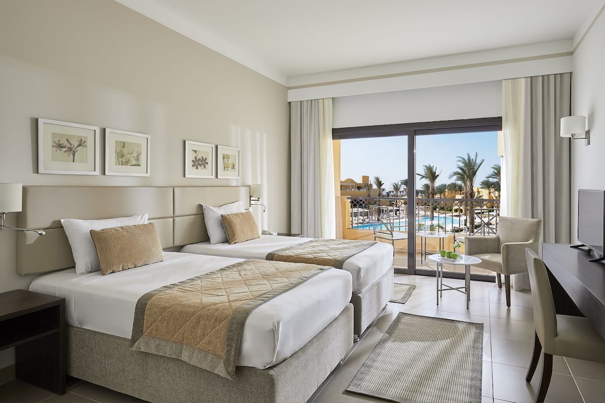 A hotel room with two beds, a desk, a chair, and a view of a pool and palm trees.