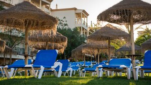 Un complexe hôtelier doté de parasols en chaume, de chaises longues bleues et d’un bâtiment avec des balcons.