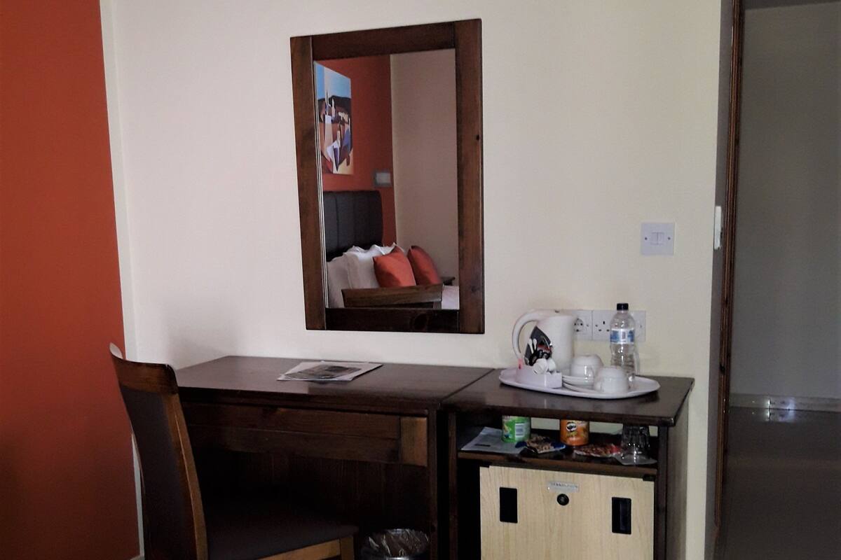 A hotel room with a bed, a desk with a chair, a small kitchenette, a trash bin, and a mirror on the wall.