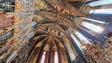 An ornate cathedral interior with intricate ceiling designs, large stained glass windows, and a prominent chandelier.