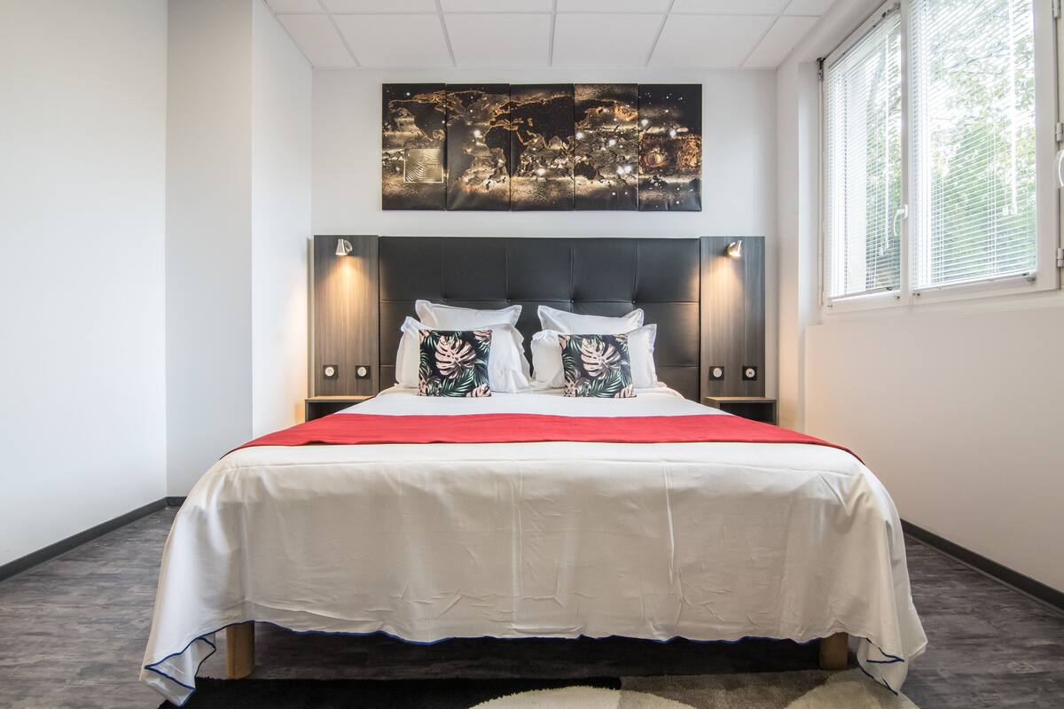 A modern bedroom with a large bed, a cityscape artwork above the bed, and a window with blinds.