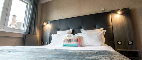 A hotel room with a large bed, a headboard with integrated lighting, a window with curtains, and a view of a building outside.