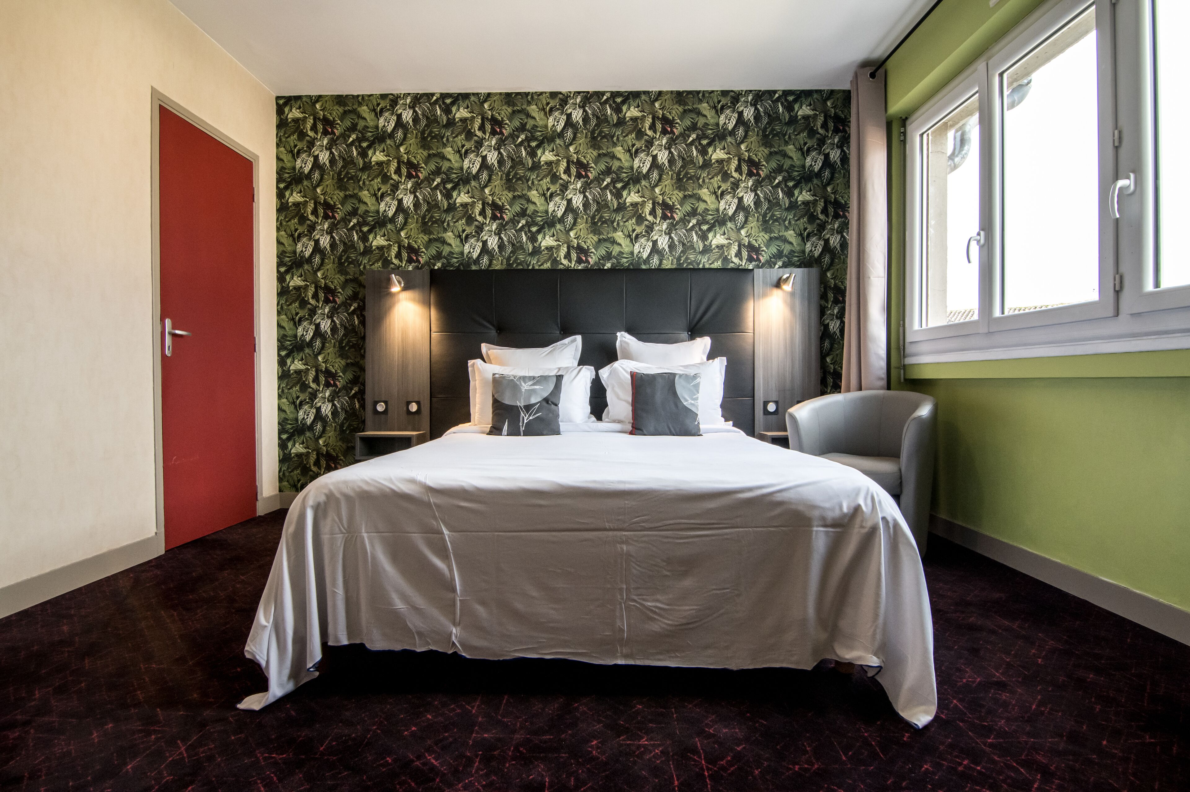 Una camera d'albergo con un letto grande, una porta rossa, una parete verde decorata con un motivo a foglie e una finestra con cornici bianche.