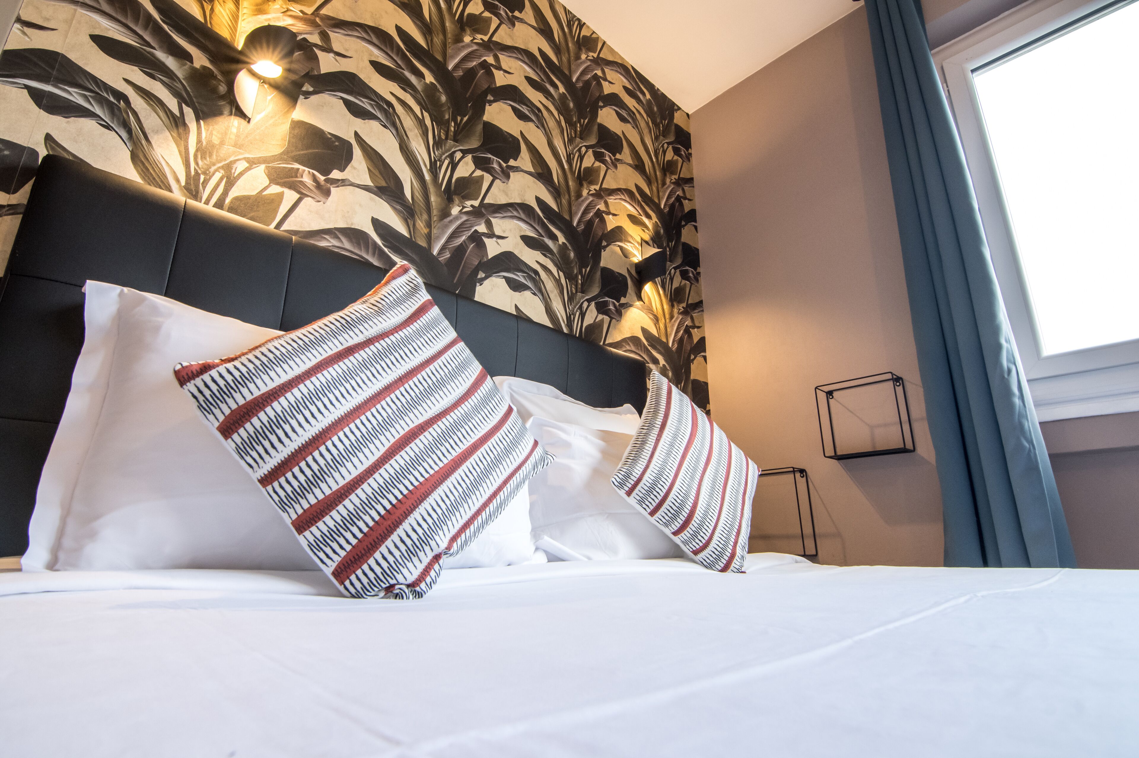 A neatly made bed with white linens and striped pillows, a dark headboard, and a floral wallpaper pattern on the wall.
