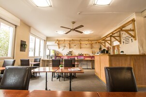 Un comedor con mesas y sillas de madera, un ventilador de techo y una cocina al fondo.