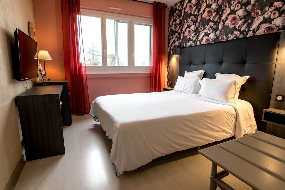 A hotel room with a bed, a TV, a desk, and a window with curtains.