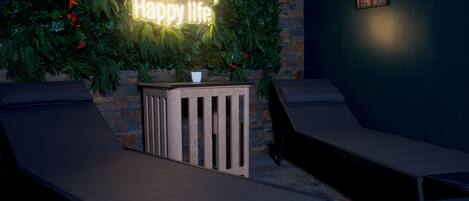 A lounge area with two reclining chairs, a neon sign reading "happy wife, happy life," and a brick wall with greenery and a framed picture.
