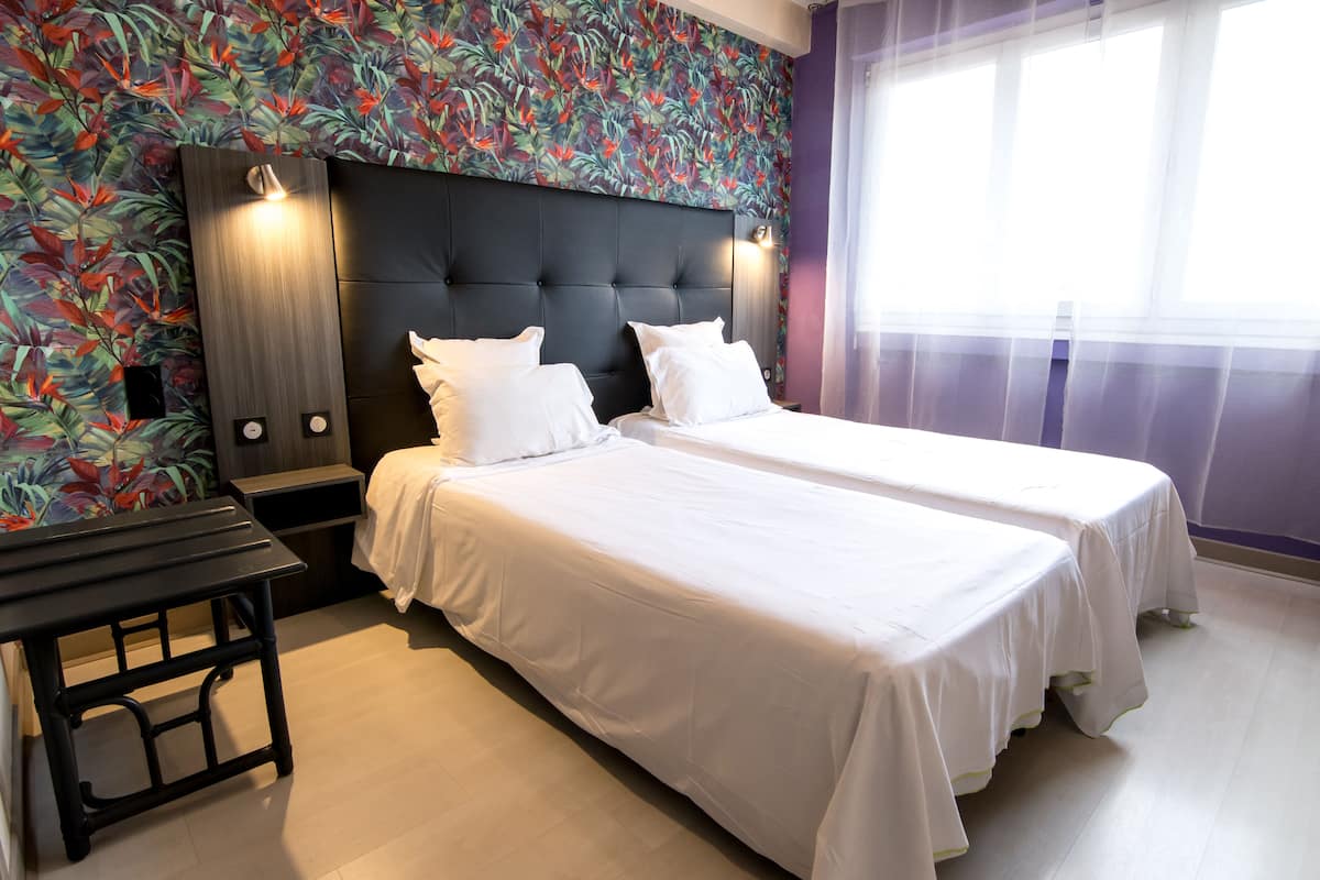 A hotel room with two beds, a wooden headboard, a small table, and a floral wallpaper.