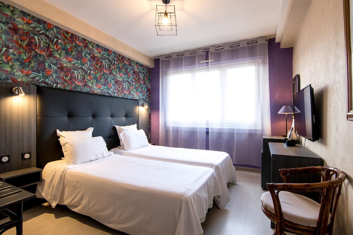 A hotel room with two beds, a large window with sheer curtains, a flat-screen TV, and a wall-mounted lamp.