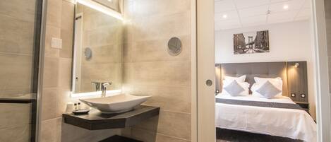Un baño moderno con un lavamanos grande, un espejo y un taburete pequeño. El baño da a un dormitorio con una cama, almohadas y una mesita de noche.