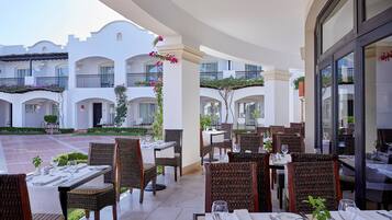 Un espace extérieur aménagé pour les repas, avec des tables et des chaises, une cour intérieure bordée d’un bâtiment blanc, et une vue sur le ciel.
