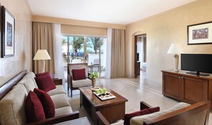 A hotel room with a sofa, armchair, coffee table with snacks, a TV, and a balcony with a view of palm trees. - JAZ Dahabeya (Dahab)