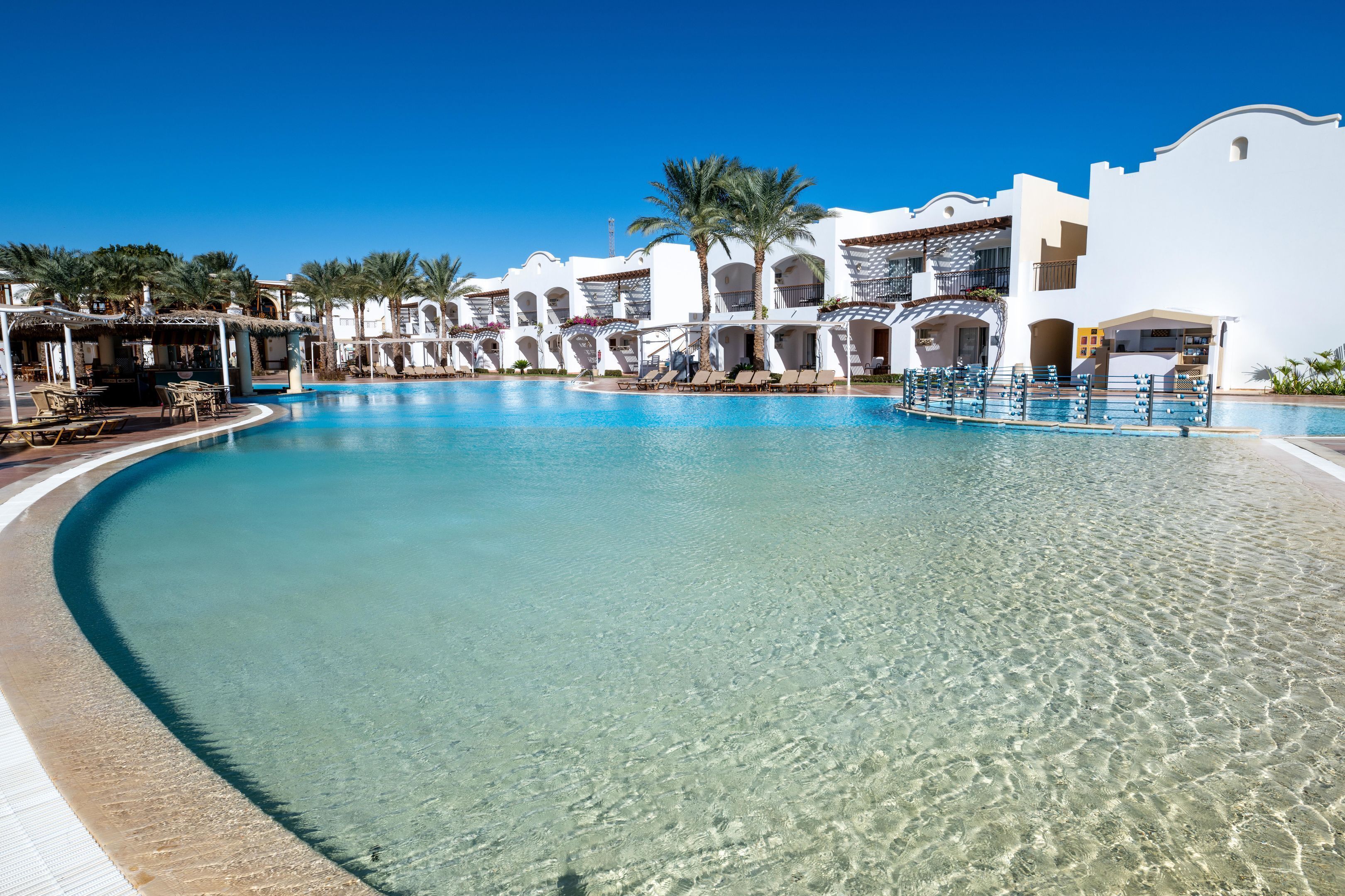 Una piscina de complejo turístico con agua cristalina, rodeada de edificios blancos con balcones y palmeras.