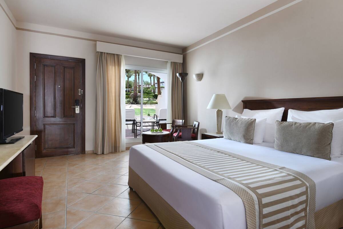 A hotel room with a bed, a desk with a television, a chair, a lamp, and a view of the outdoors.