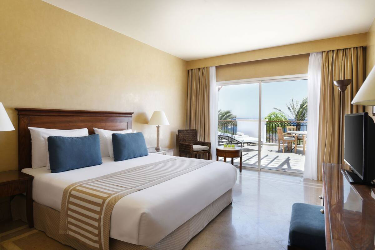 A hotel room with a large bed, a TV, a balcony with outdoor furniture, and a view of the sea.