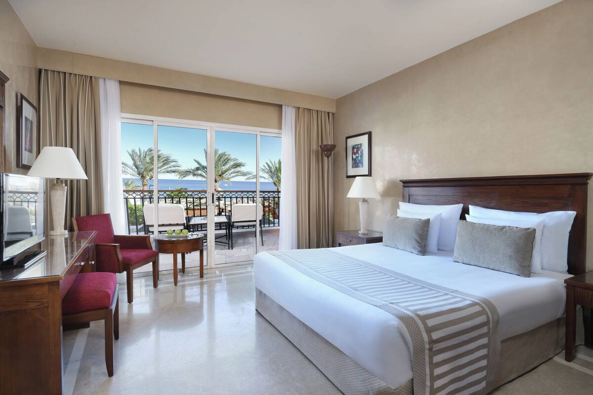 A hotel room with a large bed, a desk with a lamp, a chair, and a view of the outdoors.