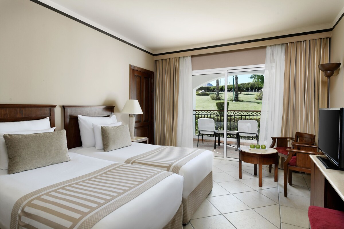 A hotel room with two beds, a TV, a small table with apples, and a balcony with chairs and a view of a garden.