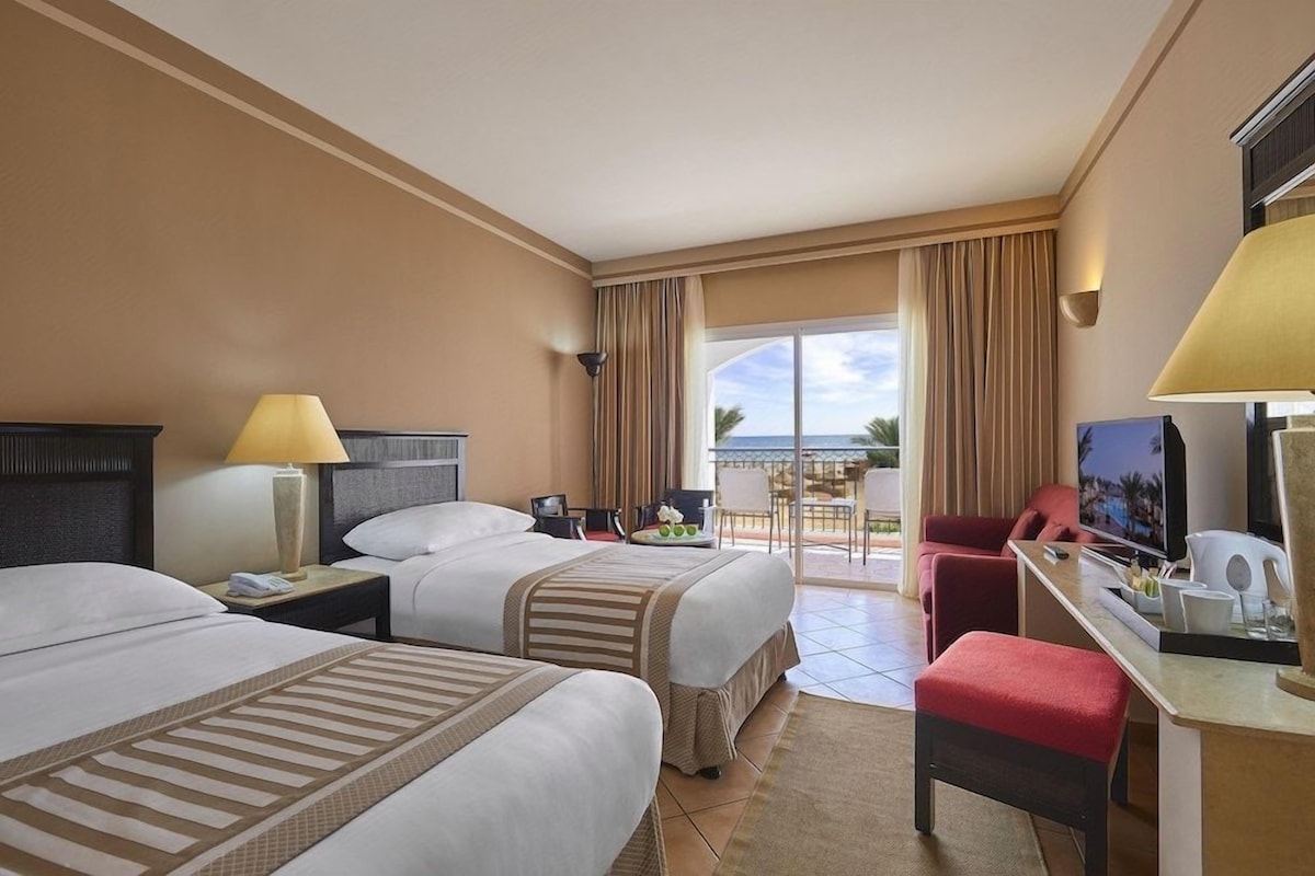 Quarto de hotel com duas camas, uma escrivaninha, televisão e varanda com vista.