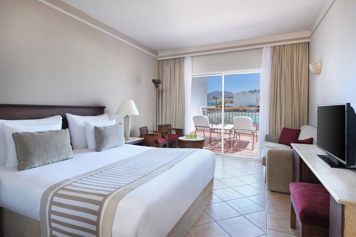 A hotel room with a large bed, a TV, a desk, and a balcony with a view of the sea.
