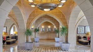 Eine geräumige Lobby mit hoher Decke, rundbogigen Steinpfeilern und einer zentralen Rezeption. Üppige Pflanzen und Sitzbereiche mit Sofas und Sesseln sorgen für Komfort.