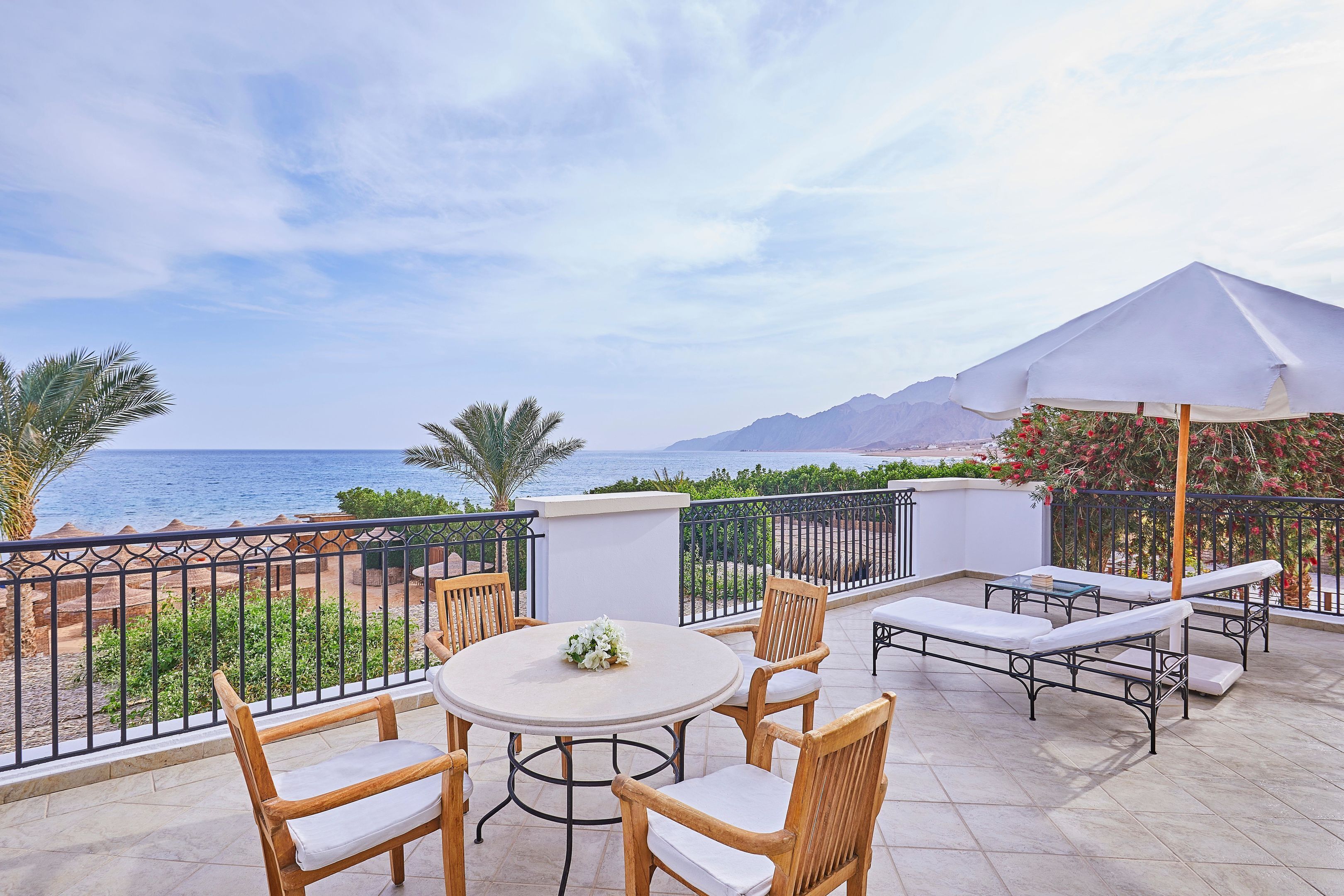 A terrace with a round table and chairs, a white umbrella, and a view of the sea and mountains.