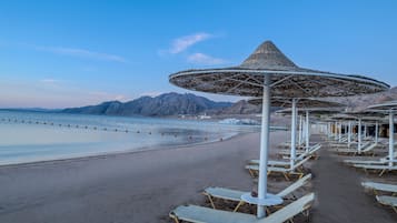 Una playa con filas de tumbonas y sombrillas, cielo despejado y montañas al fondo.