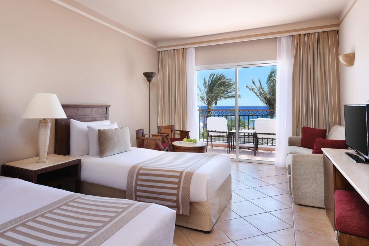 A hotel room with two beds, a desk, a chair, a TV, and a balcony with a view of the sea.