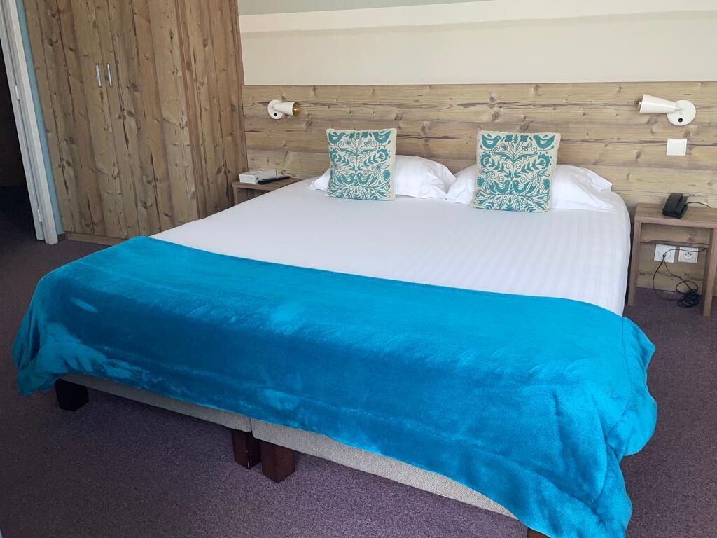 Una cama con colcha azul, dos almohadas decorativas, cabecera de madera, mesita de noche con lámpara y toma de corriente.
