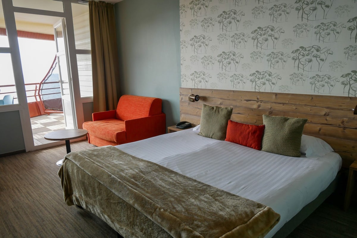 A hotel room with a bed, a chair, a small table, and a view of the sea through a sliding door.