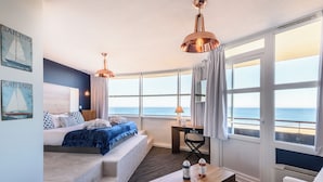 A room with a bed, a desk, a chair, a coffee table, and large windows with a view of the sea.