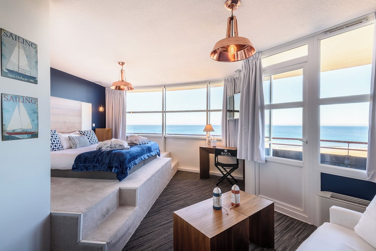 A room with a bed, a desk, a chair, a coffee table, and large windows with a view of the sea.