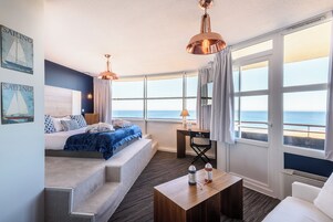 A room with a bed, a desk, a chair, a coffee table, and large windows with a view of the sea.