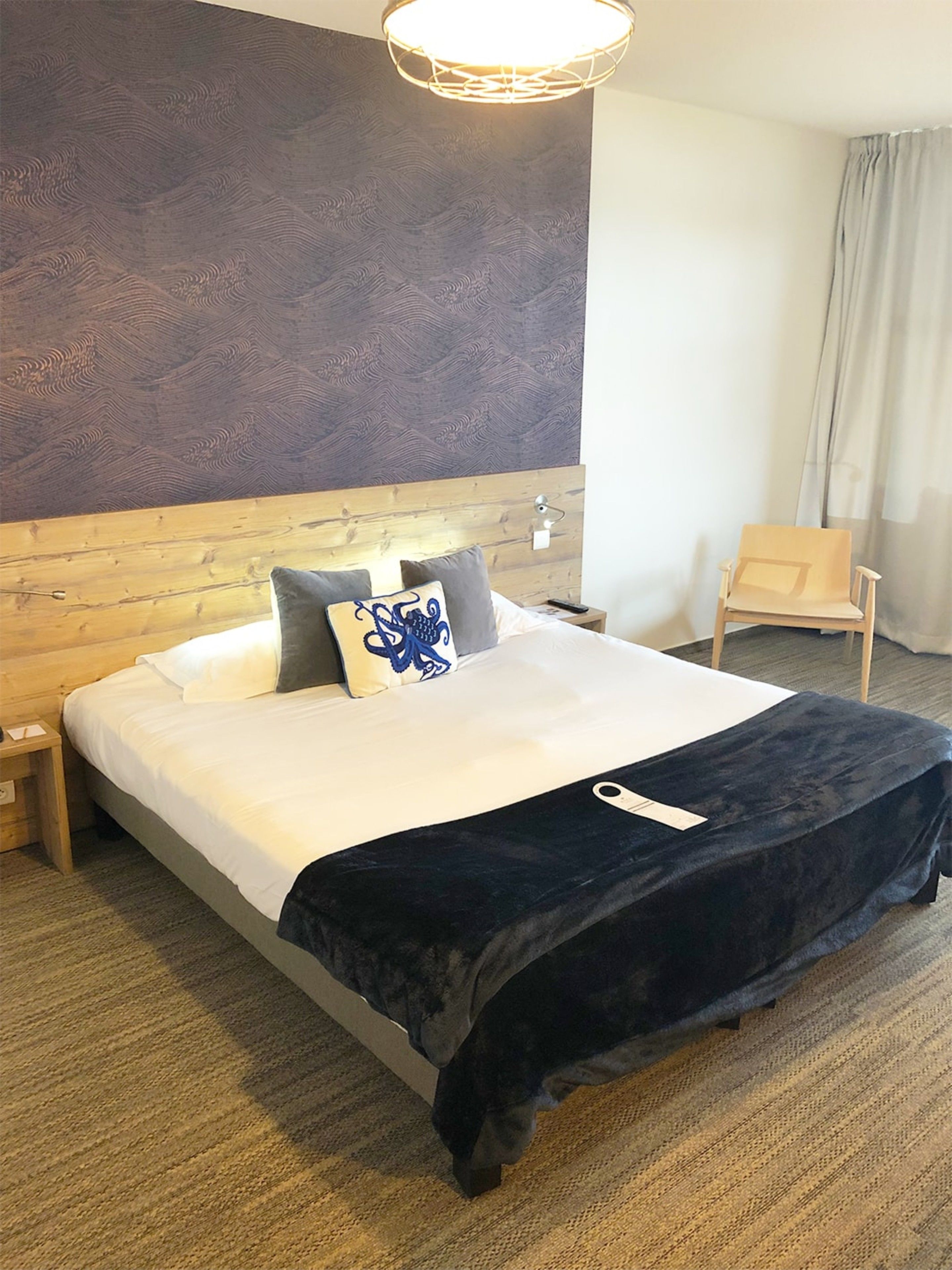 A hotel room with a wooden headboard, a bed with white linens, a dark blue throw blanket, and a single pillow with a blue design.