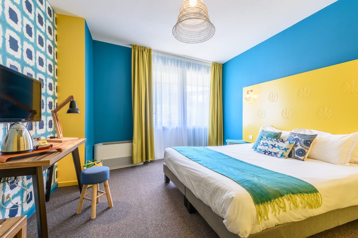 A hotel room with a bed, a desk with a lamp, a television, and a window with curtains.