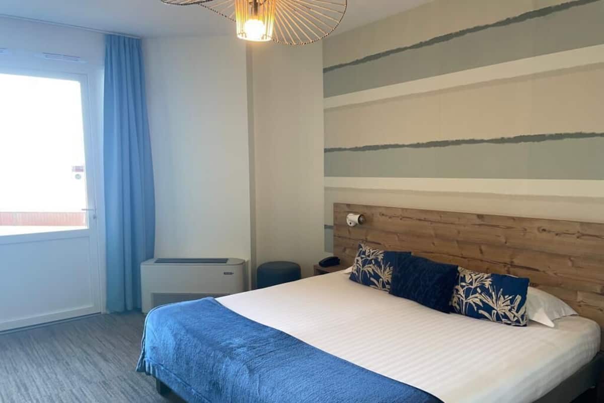 A hotel room with a large bed, a wooden headboard, a window with curtains, and a ceiling light fixture.