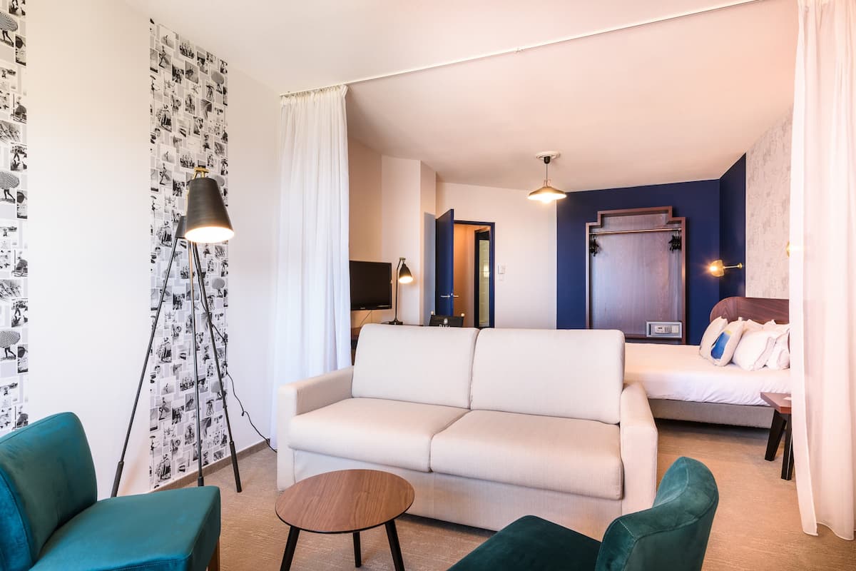 A modern hotel room with a white sofa, teal armchairs, a small wooden side table, and a large mirror.