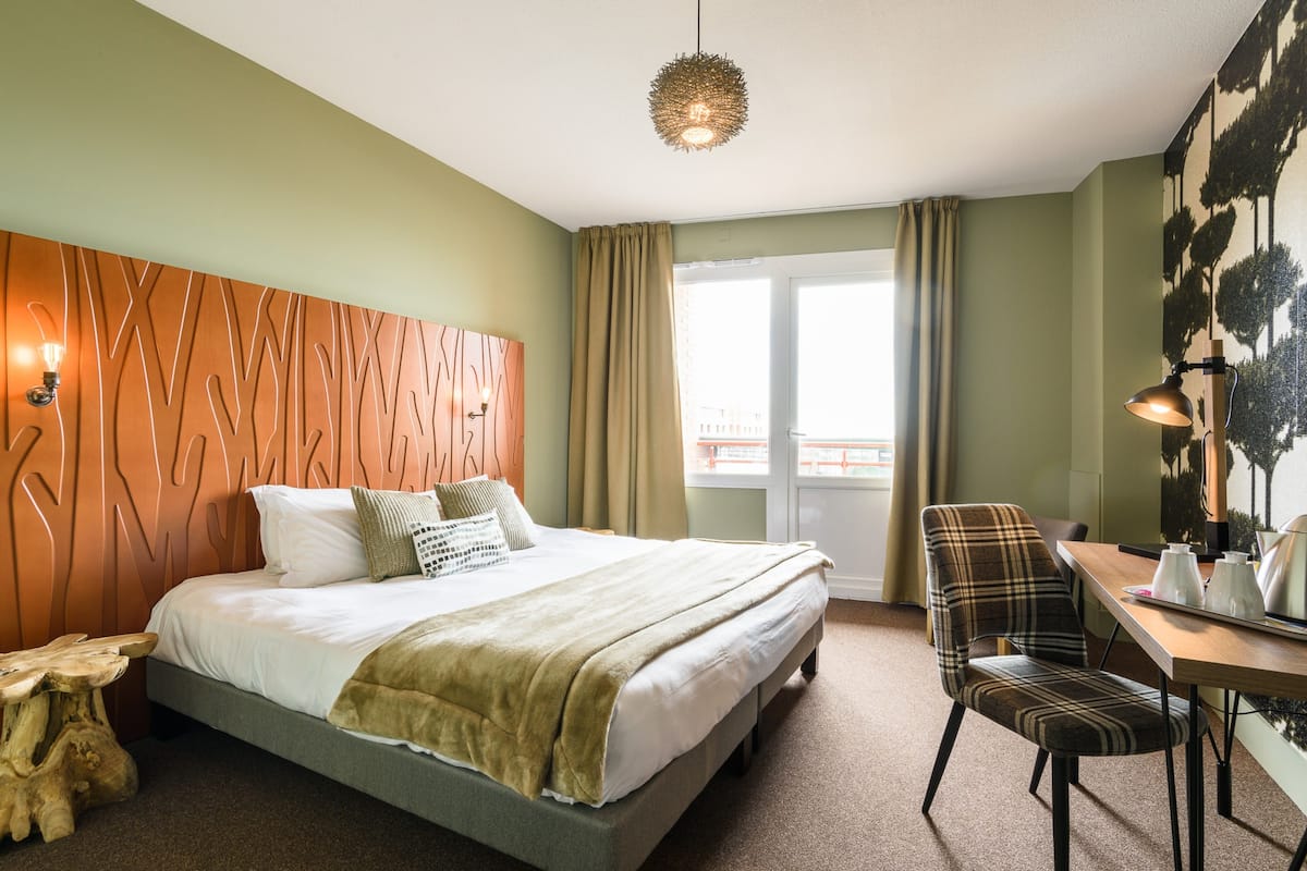 A hotel room with a bed, a desk, a chair, a wooden headboard, a window with curtains, and a wall-mounted light fixture.