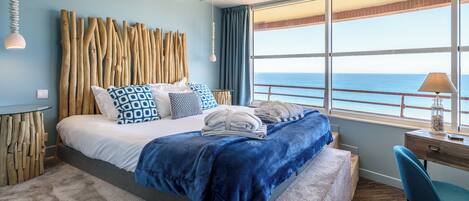 A hotel room with a bed, a desk, a chair, a view of the sea, and a unique wooden headboard.
