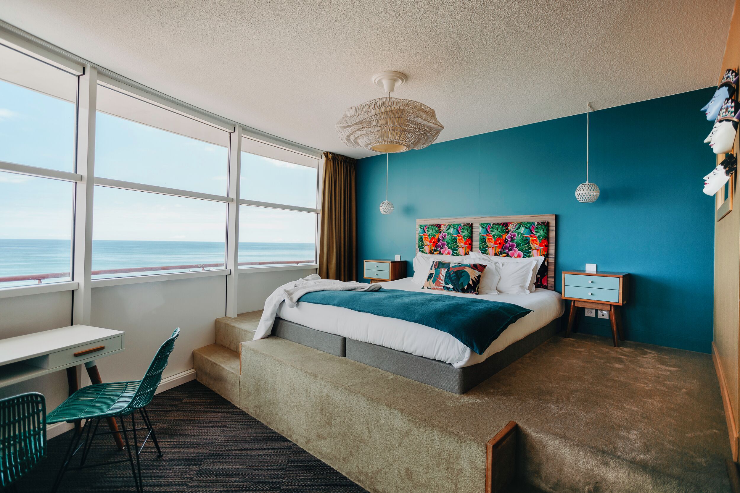 Un dormitorio con cama, escritorio, silla y vista al mar, con una pared azul.