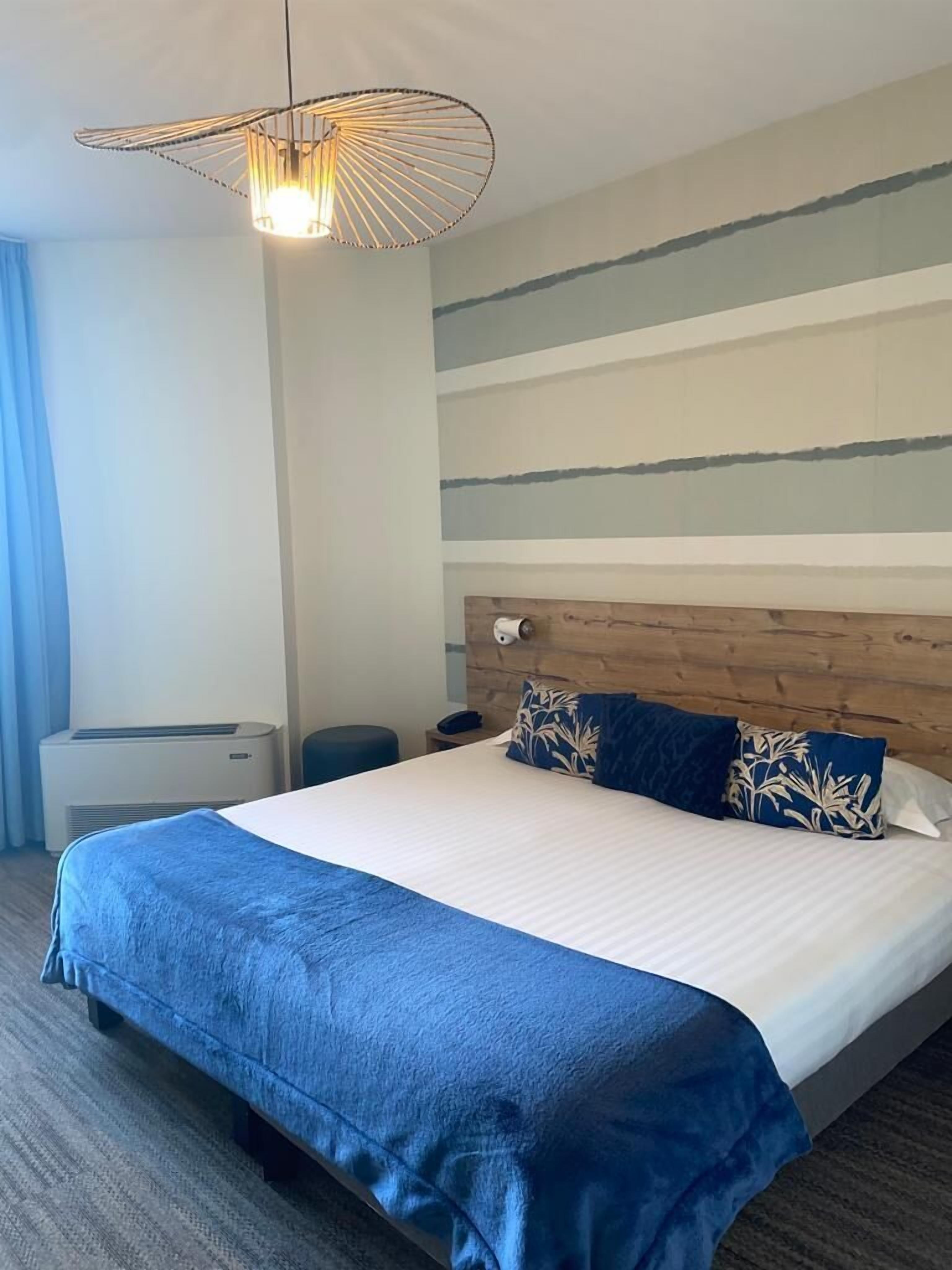 A hotel room with a large bed, a wooden headboard, a modern pendant light, a small bedside table, and a wall-mounted air conditioning unit.