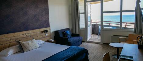 Habitación de hotel con una cama grande, un escritorio con silla, un sofá y vista al mar a través de las ventanas.