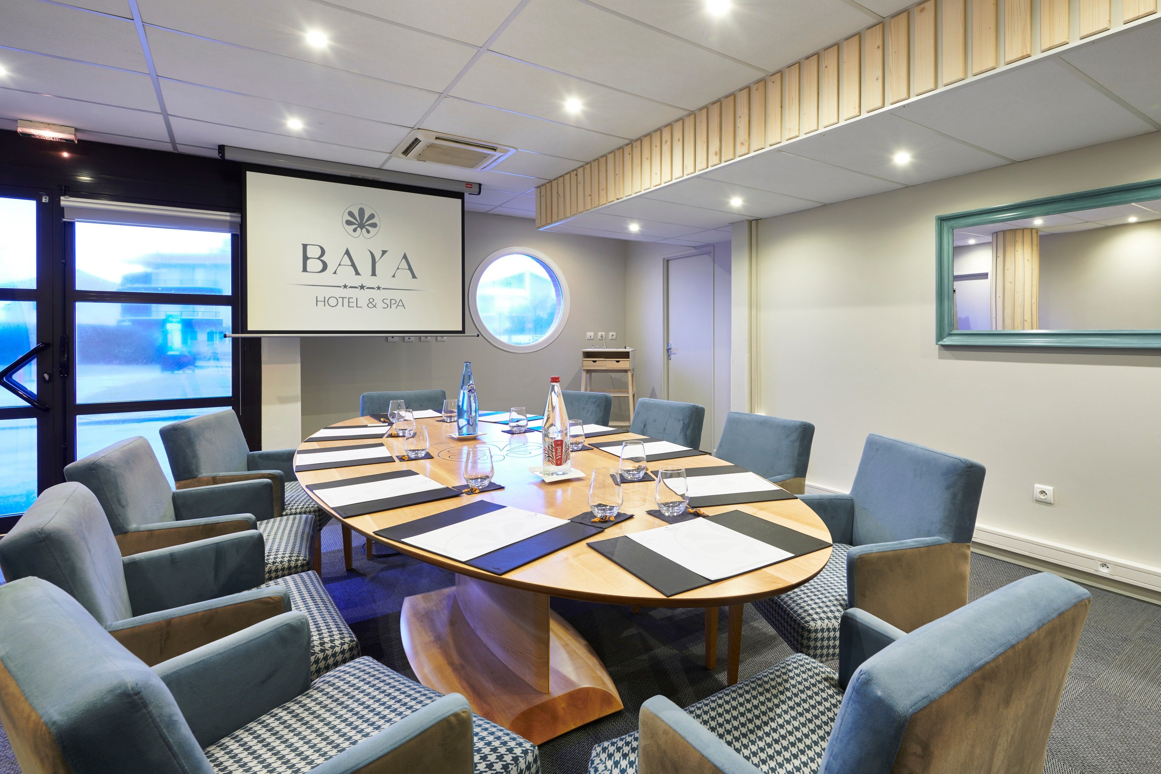 A conference room with a large round table, blue upholstered chairs, and a projector screen displaying "BAYA HOTEL & SPA".