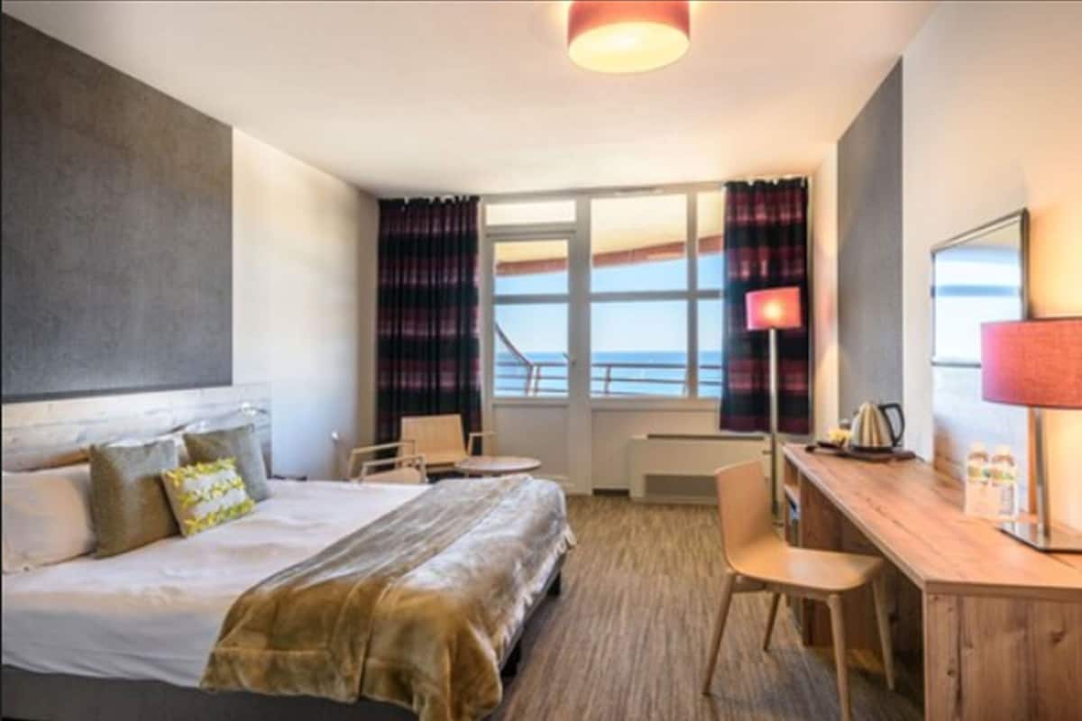 A hotel room with a bed, desk, chair, and a view of the sea.