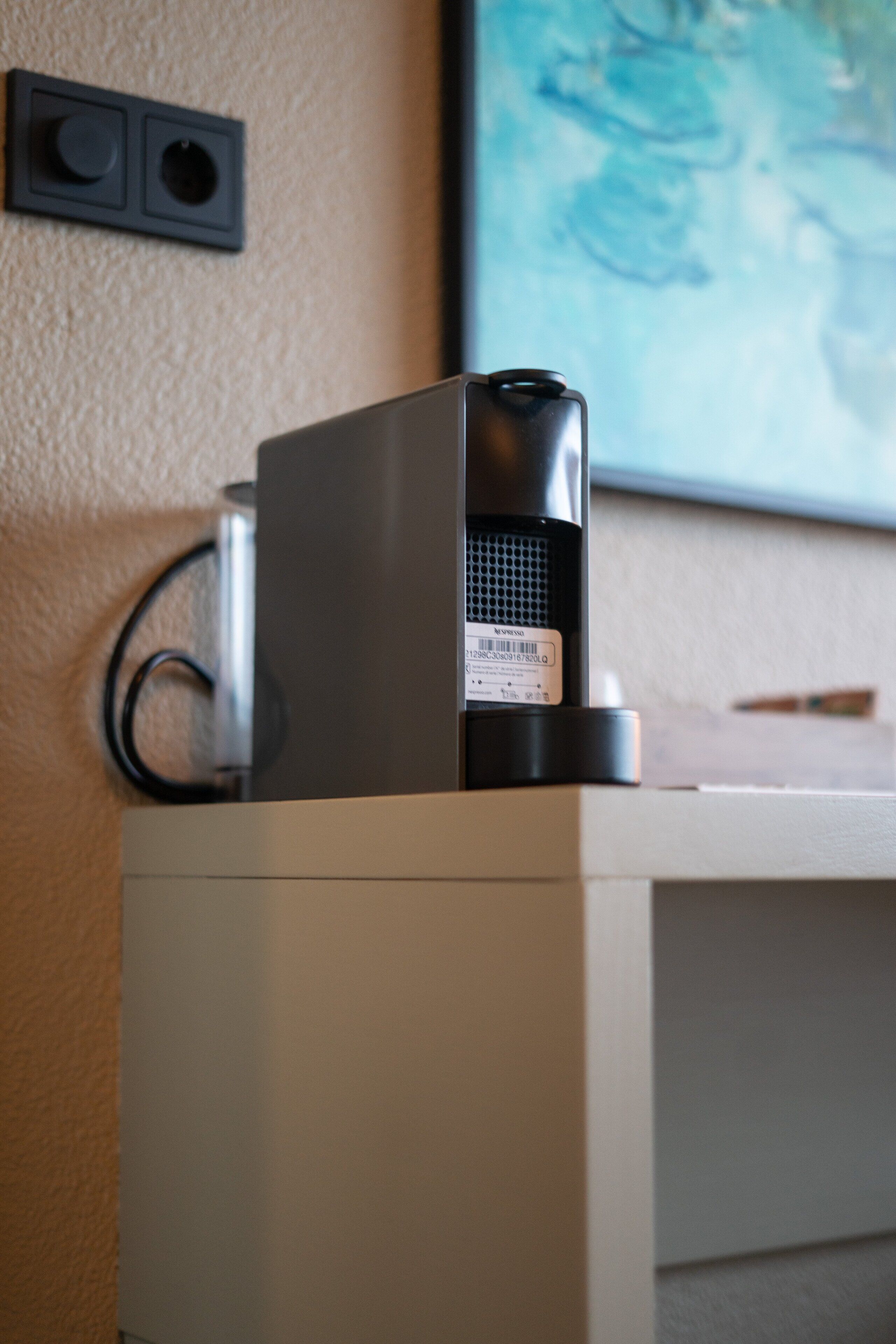 Un proyector sobre un soporte blanco frente a una pared beige, con un cuadro enmarcado encima.