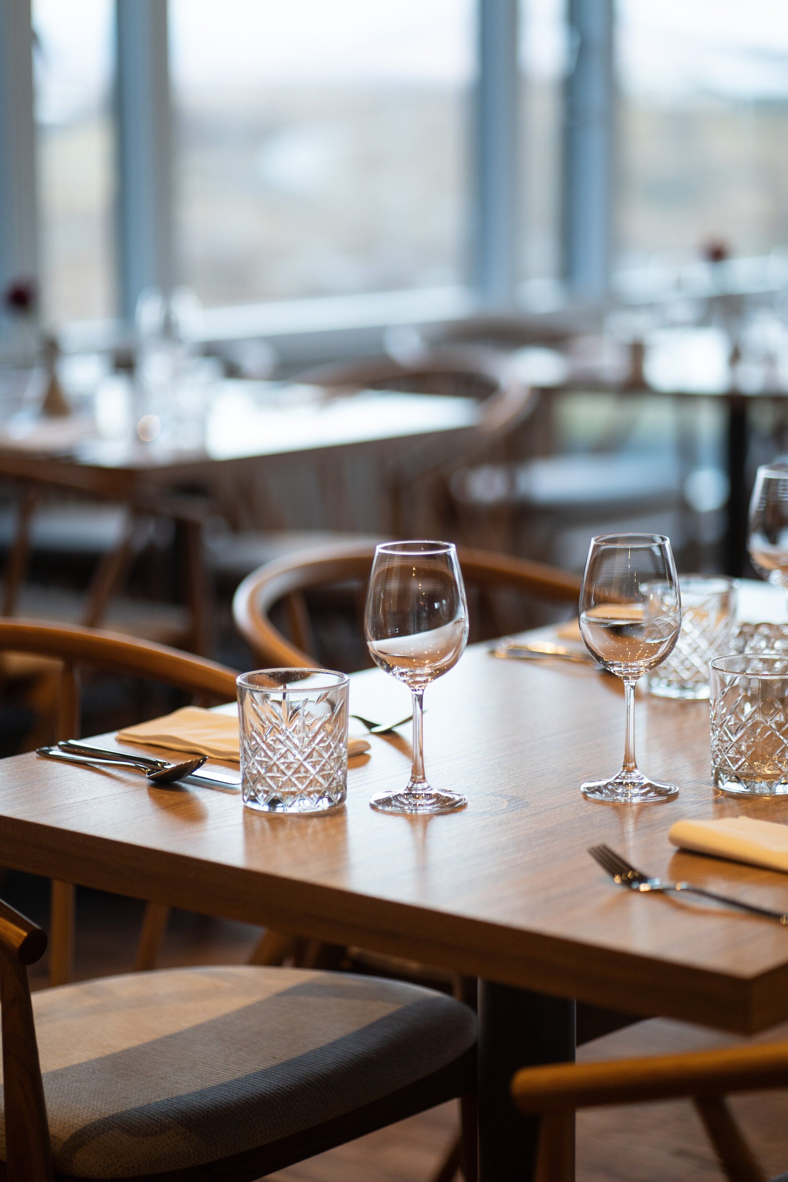 A dining table with clear glassware and cutlery, wooden chairs, and a view through large windows.
