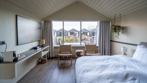 Een hotelkamer met een bed, een bureau met stoelen, een groot raam met gordijnen, een tv aan de muur en een hangplant.