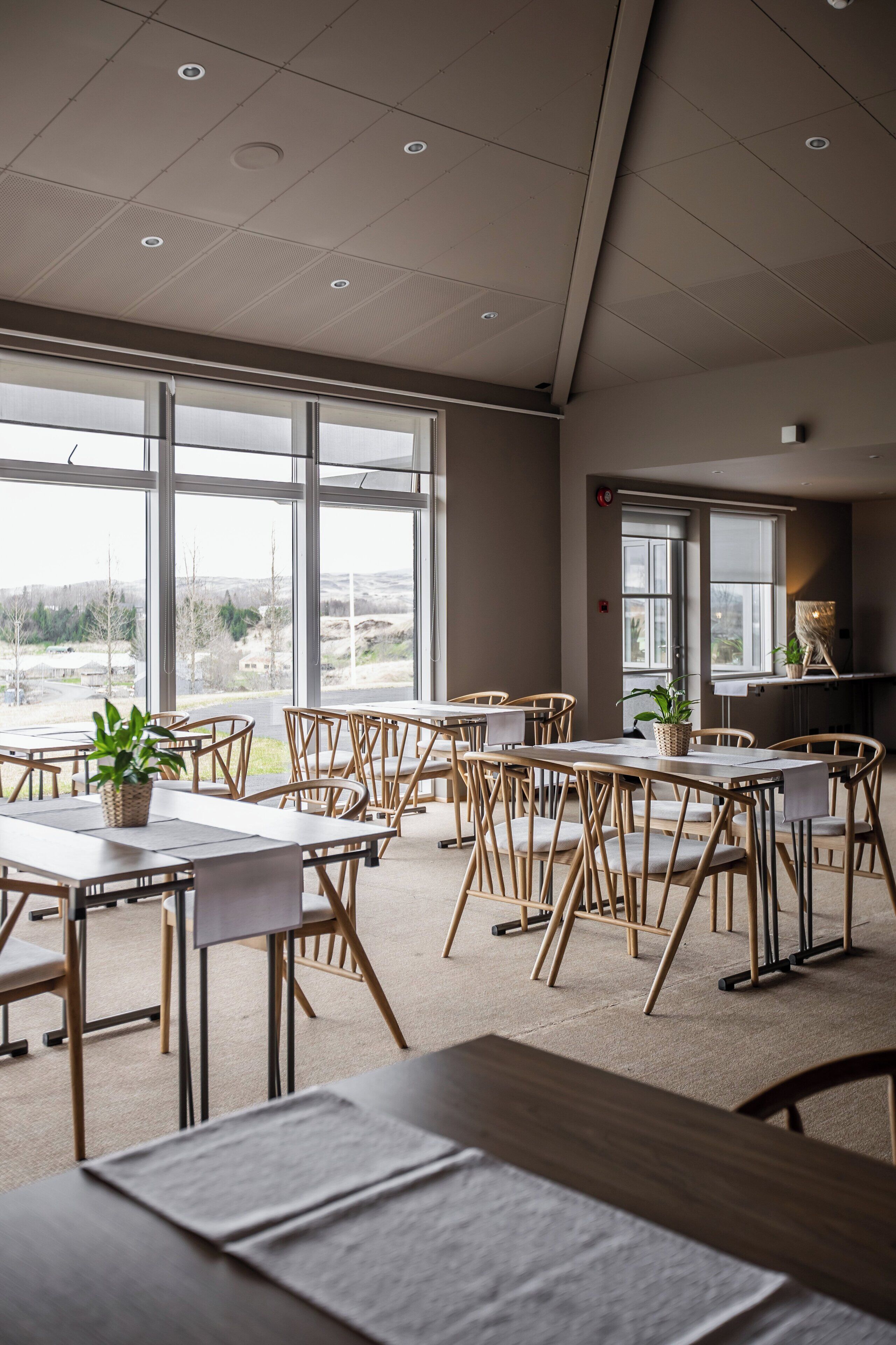 A spacious room with large windows, wooden tables, and chairs arranged for dining.